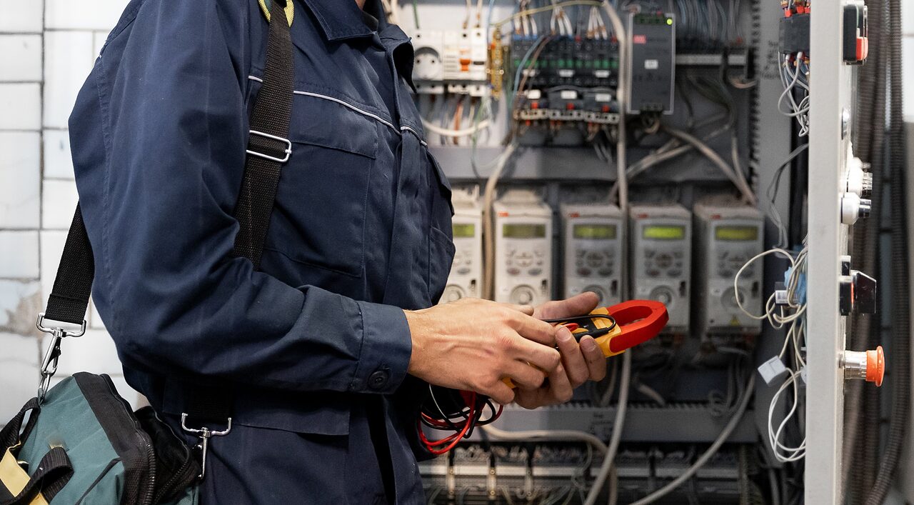 An electrician wearing a dark blue jumpsuit and carrying a tool bag examines and tests electrical wiring with a multimeter in front of an open electrical panel.
