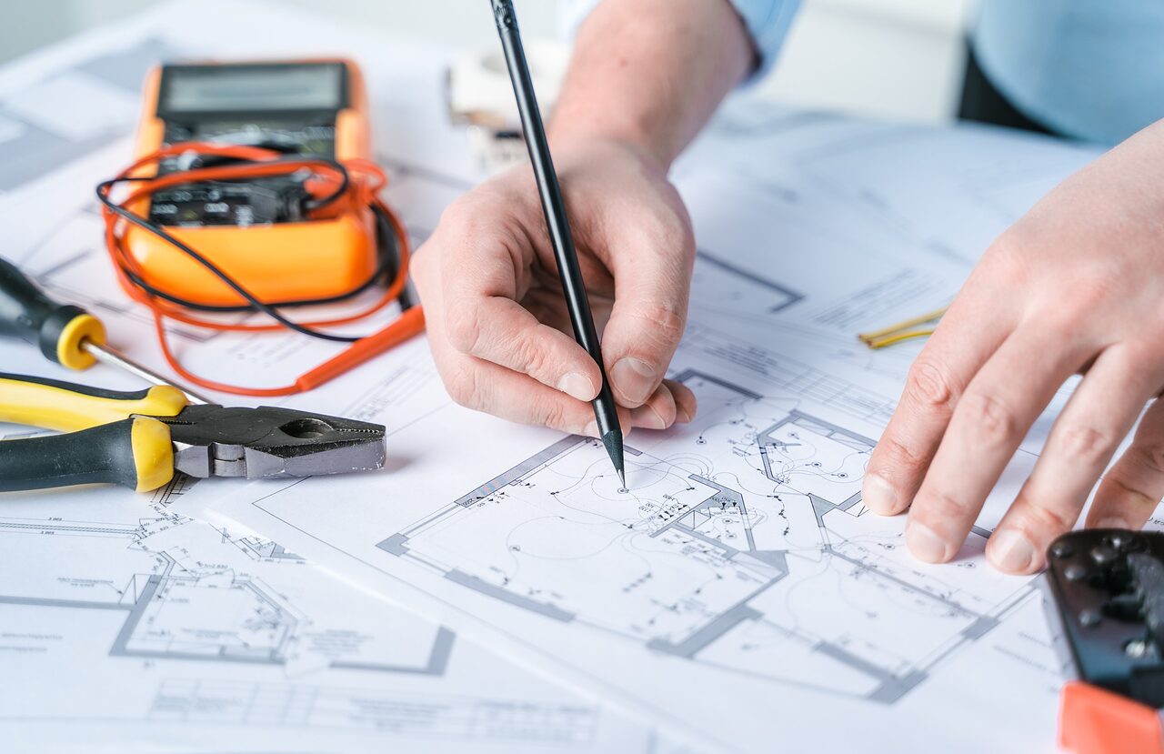 Person working on an architectural blueprint with tools including pliers, a screwdriver, and a multimeter on a desk.