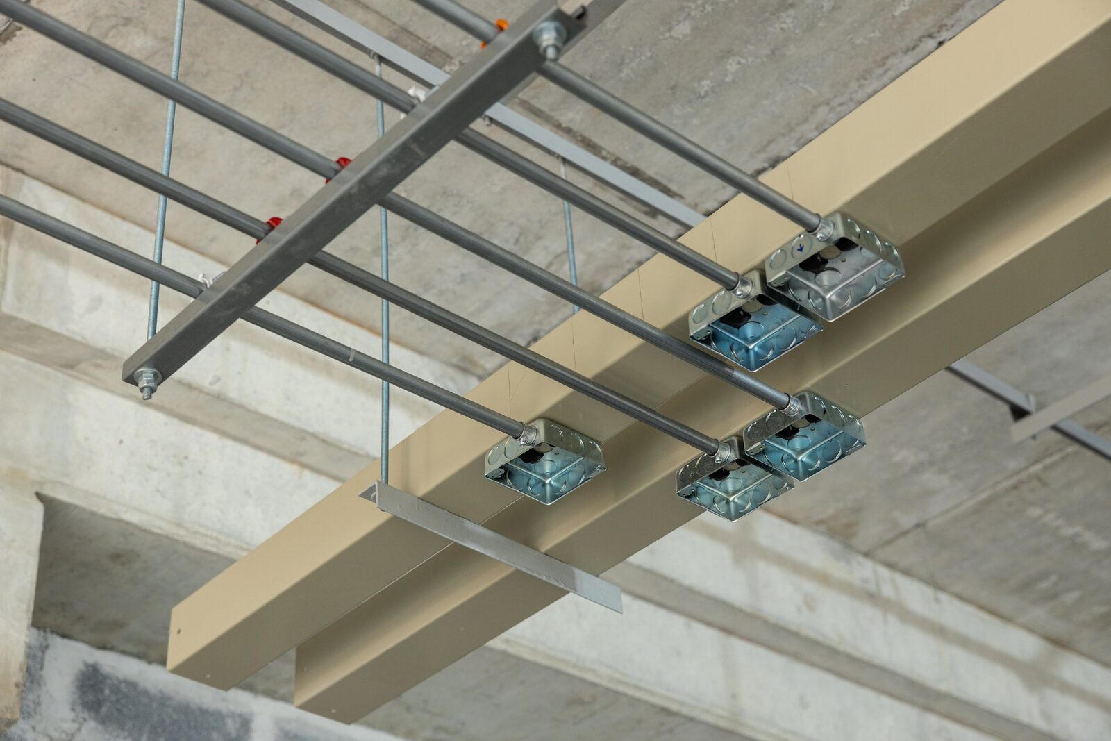 Commercial electrical rough-in below a concrete slab: steel conduit runs feeding ganged metal device boxes mounted beside a painted structural beam, with ceiling grid hangers in place.