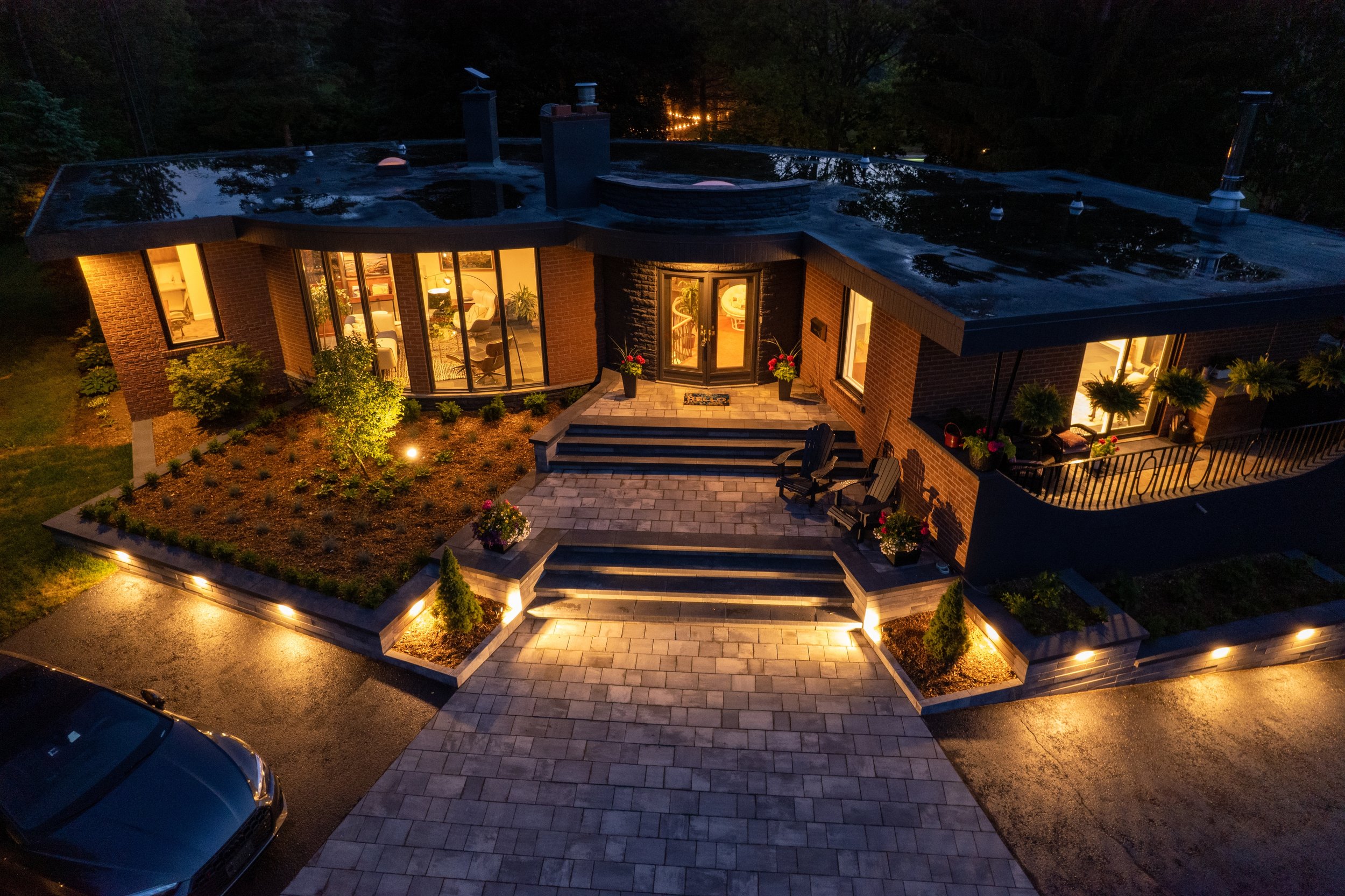 A modern, illuminated brick house at night with a paved driveway, front porch with stairs, potted plants, and outdoor seating.
