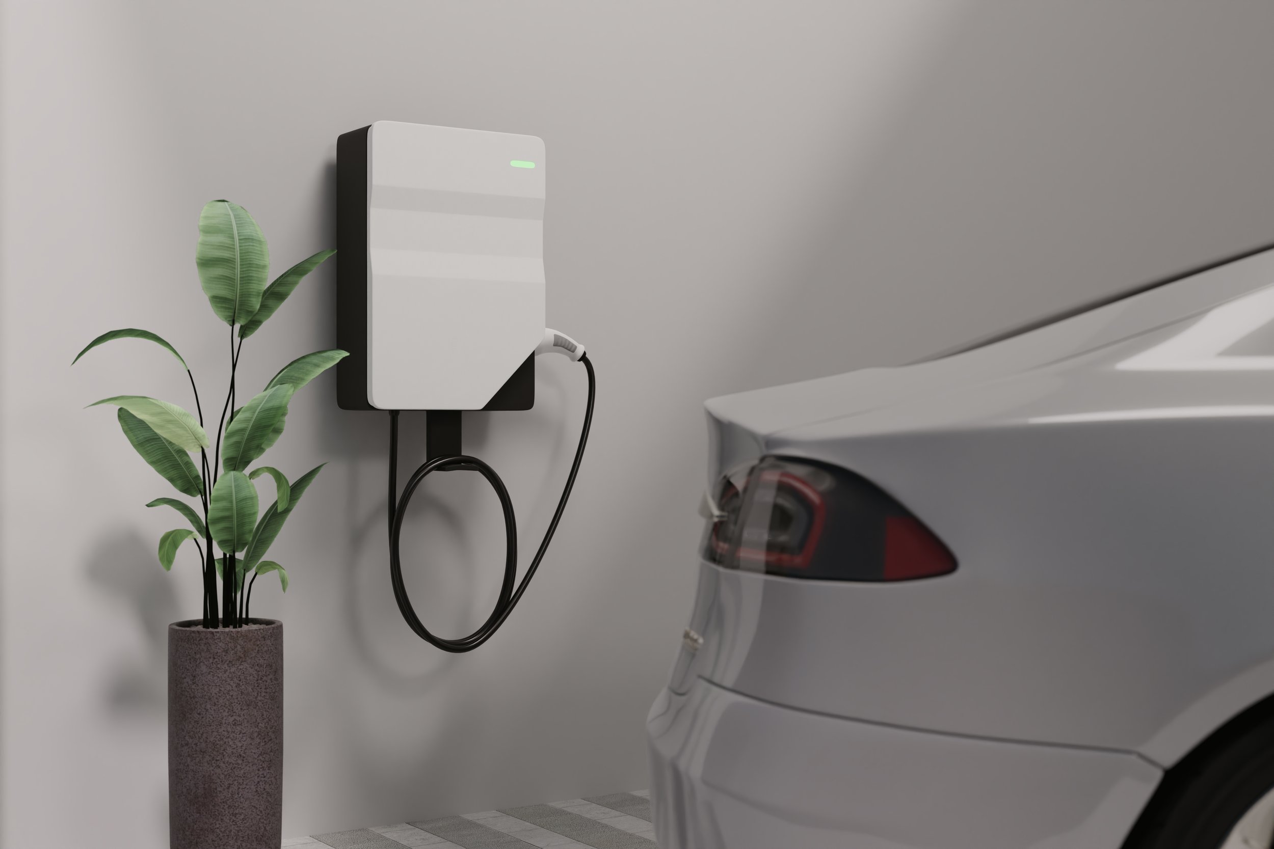 A wall-mounted EV charging unit with its cable coiled beneath it, beside a potted plant, with the rear of a light grey car parked alongside.