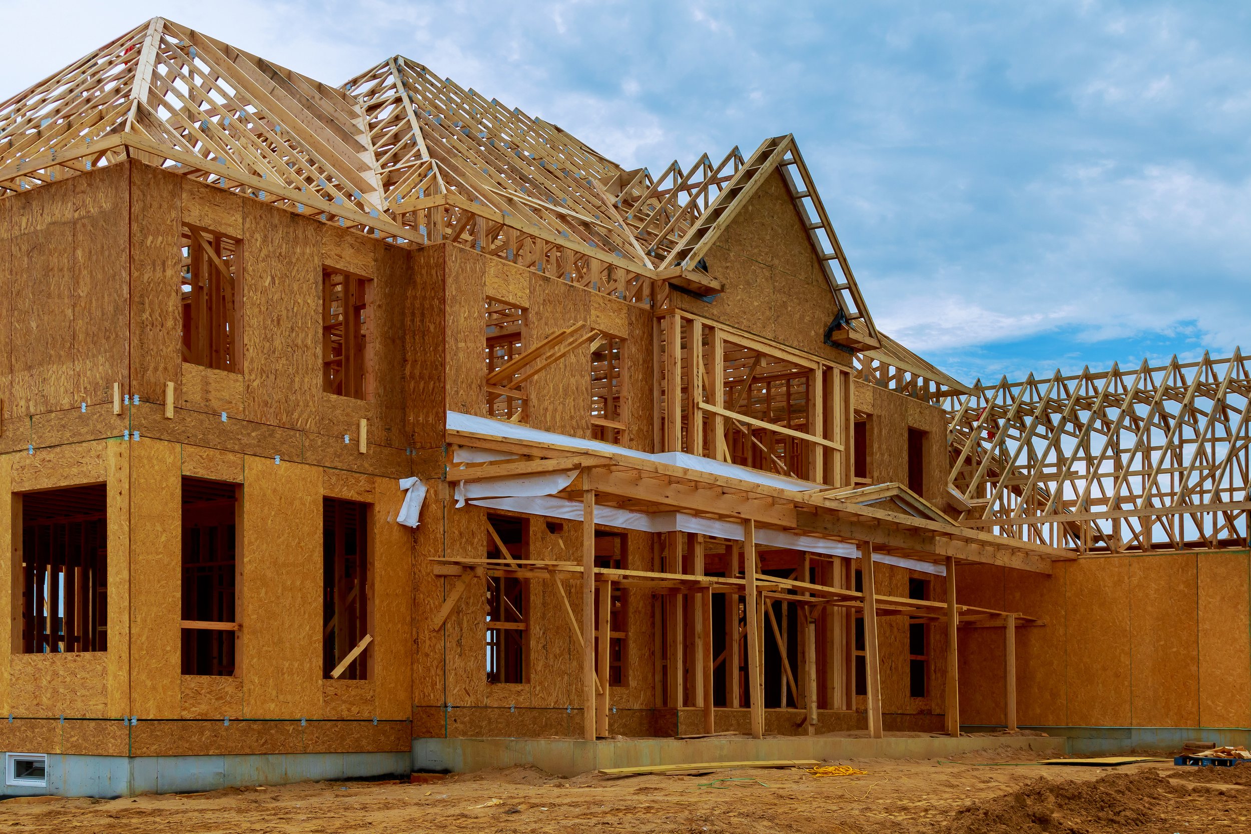 Wooden house under construction with exposed framing and roof trusses, dirt ground, and cloudy sky.