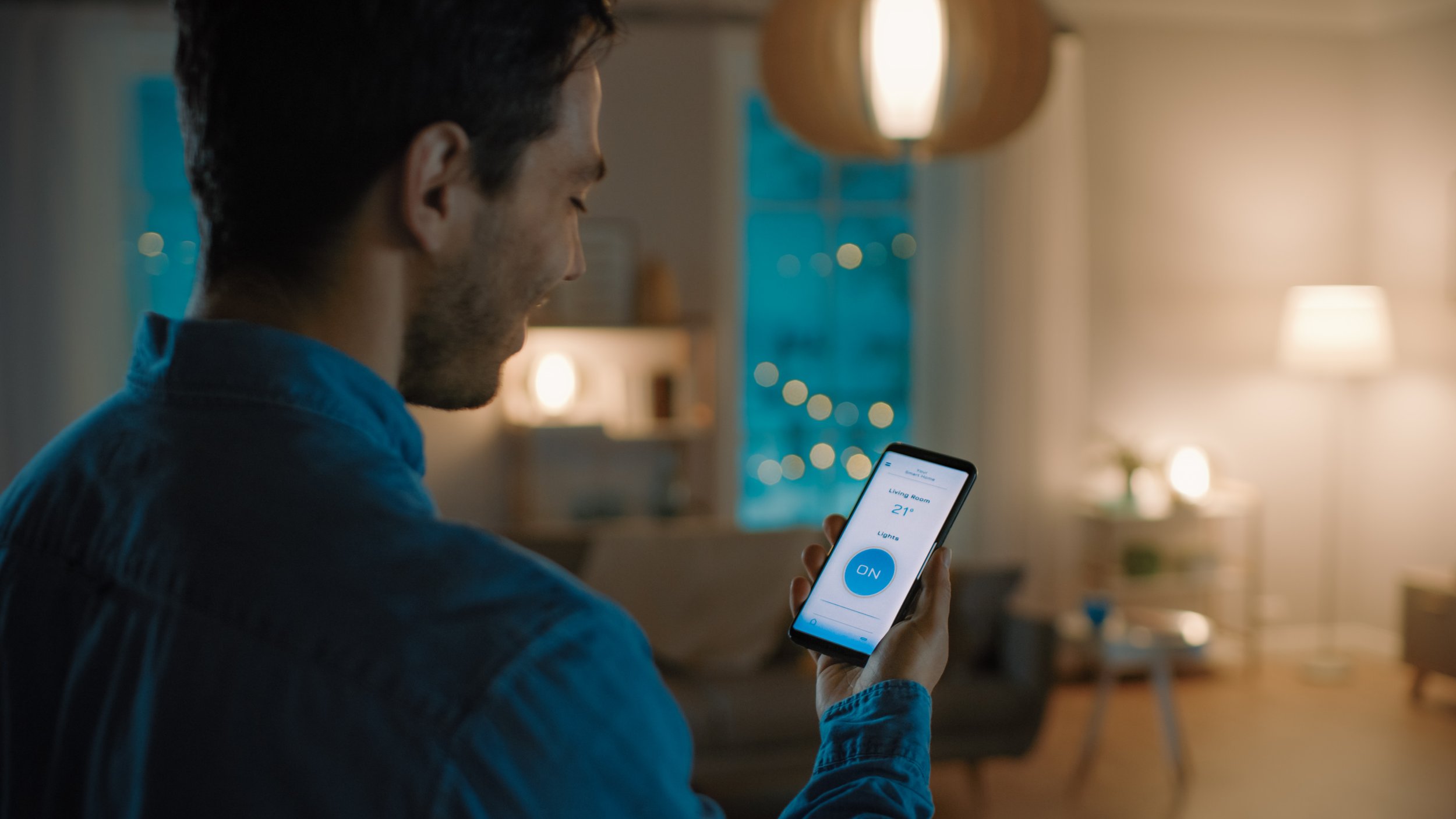 A man operates a smartphone controlling a smart home device in a cozy living room at night.