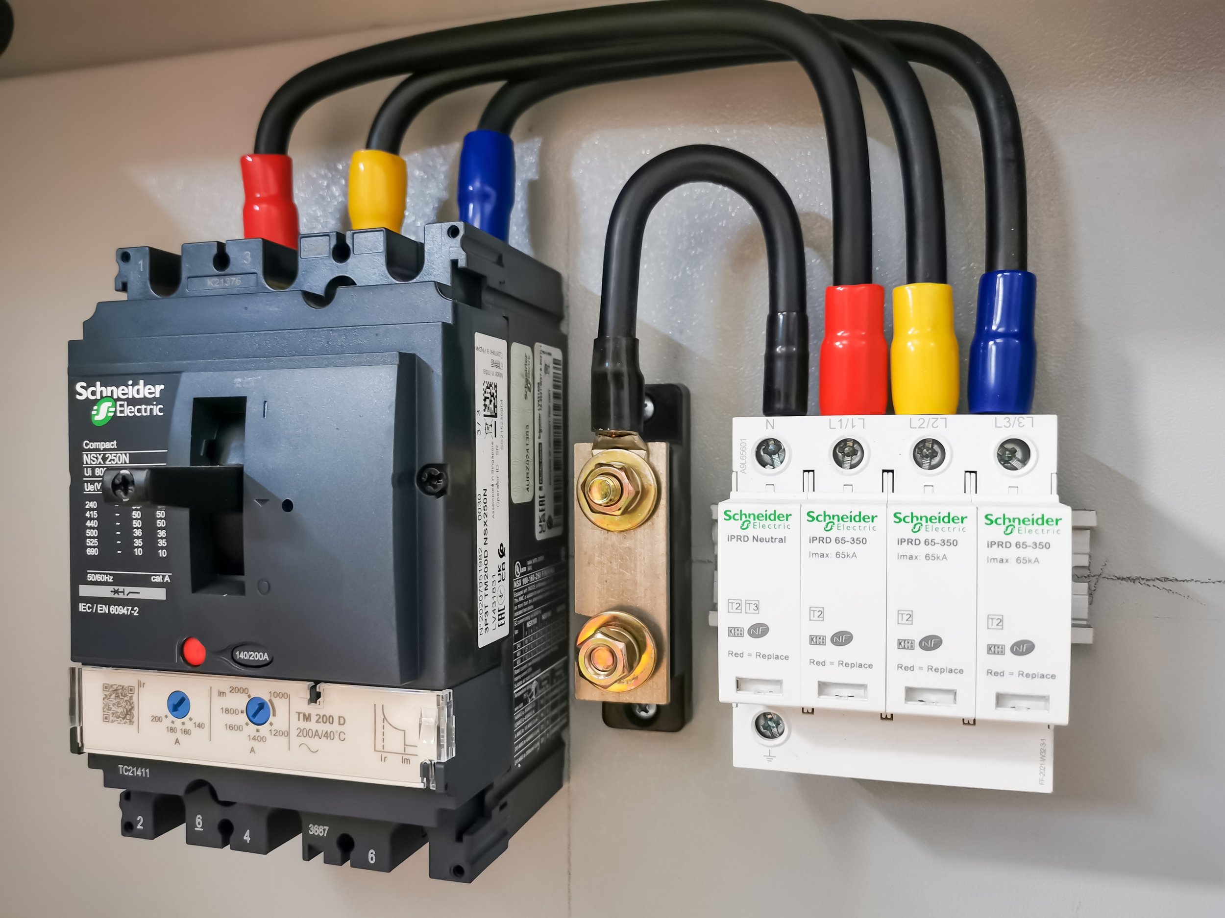 Electrical panel with a Schneider Electric circuit breaker, three white terminal blocks with colored wires connected, and a black grounding wire with a brass connector in the middle.