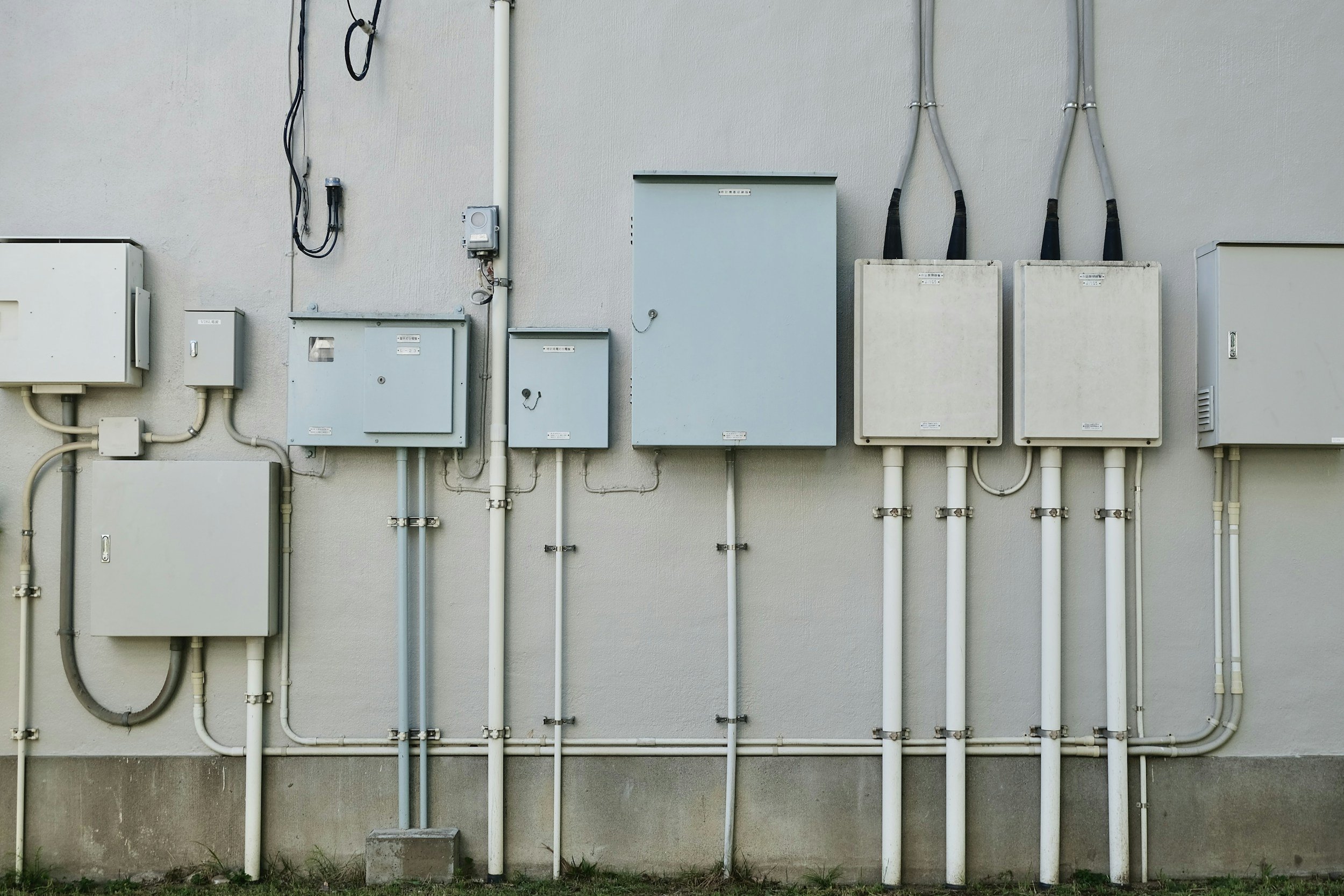 Multiple electrical and utility boxes and meters mounted on a beige exterior wall, with pipes and wires running vertically and horizontally.